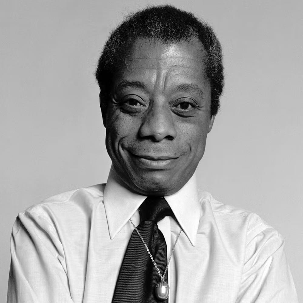 a black and white photo of the writer James Baldwin, standing in a white collared shirt and black tie