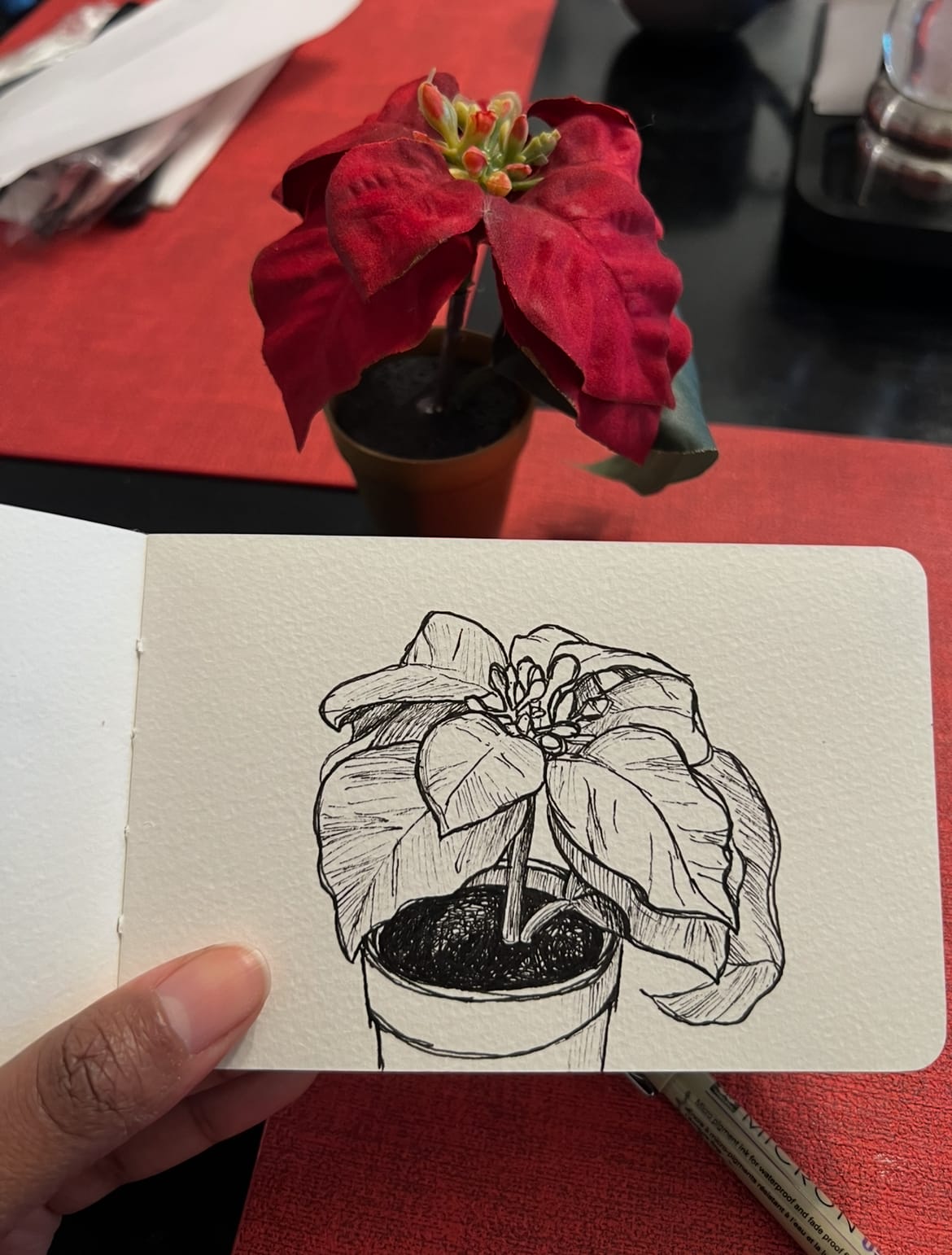 Alleanna holds a mini Moleskine sketchbook with her sketch of the mini poinsettia decoration in front of her.